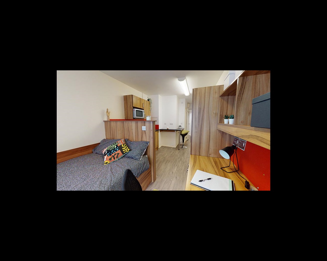 A bedroom in Cambridge's CB1 accommodation, with a bed, desk, shelves, and a kitchen area in the distance