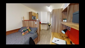 A bedroom in Cambridge's CB1 accommodation, with a bed, desk, shelves, and a kitchen area in the distance