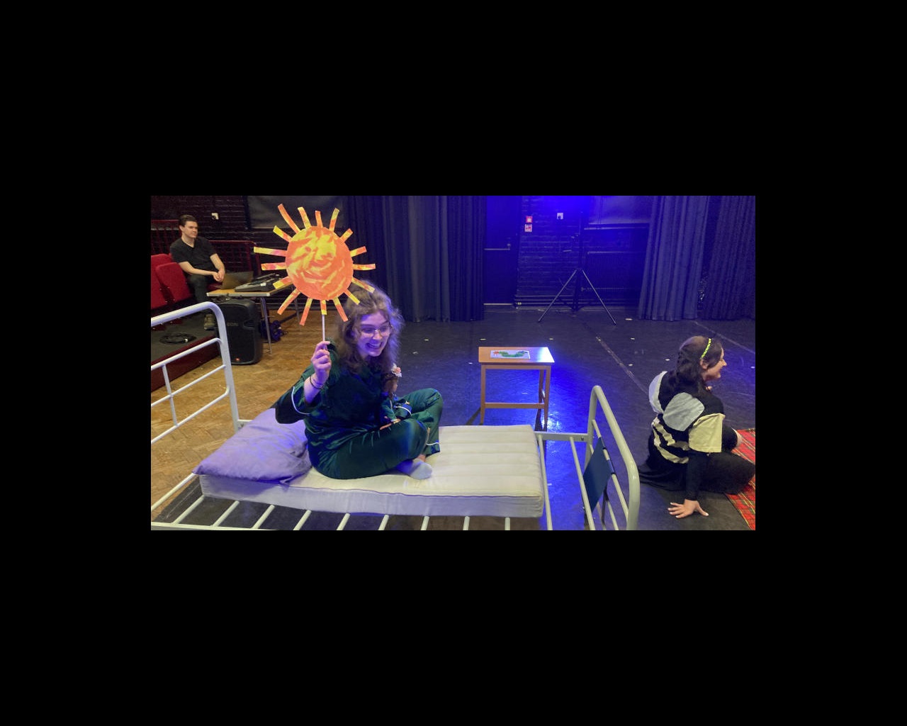 Female drama student sitting on hospital bed holding up cardboard sun with another sat in front of her on stage