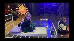 Female drama student sitting on hospital bed holding up cardboard sun with another sat in front of her on stage