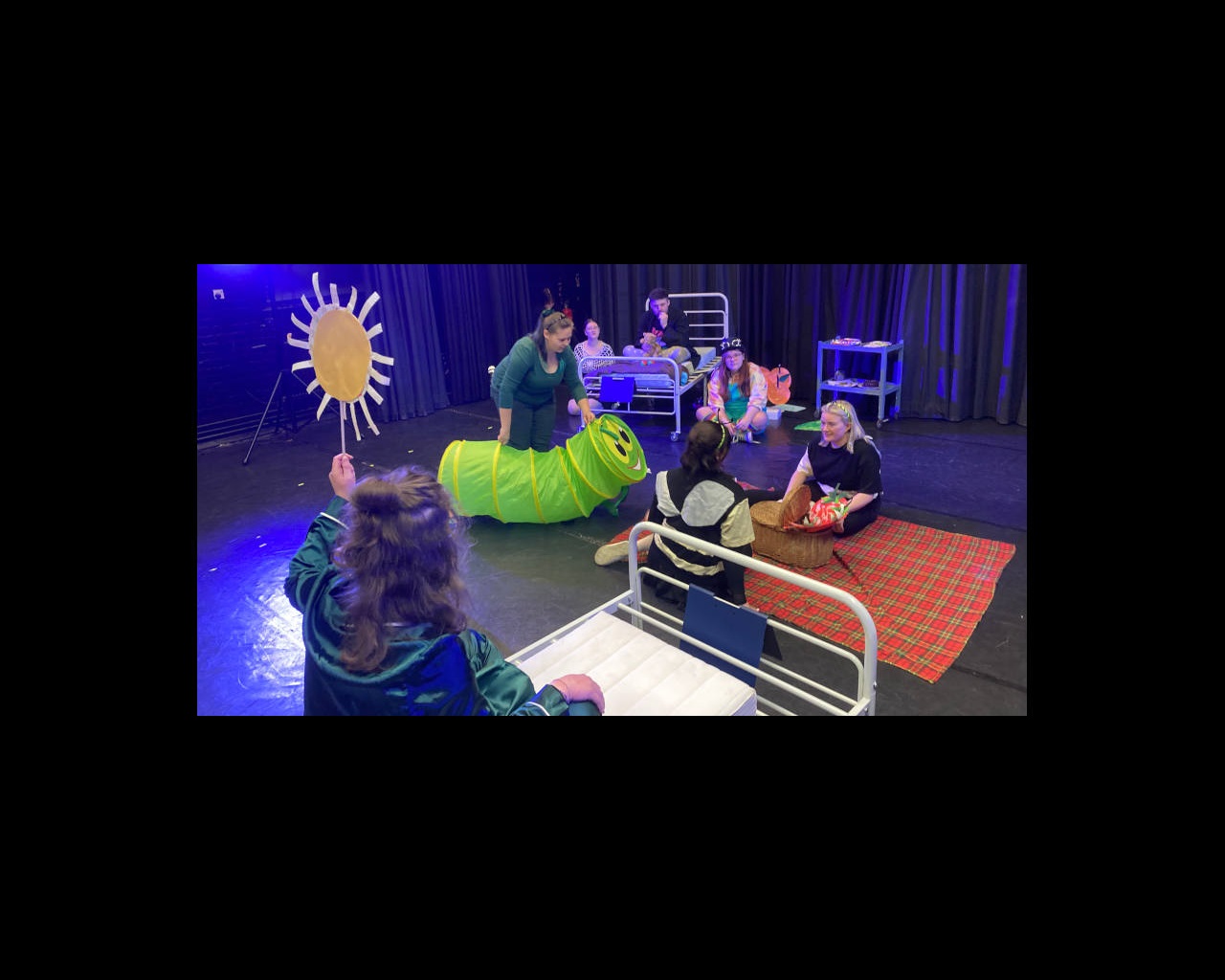 Drama students on stage in hospital set in various poses, one moving a large model Hungry Caterpillar