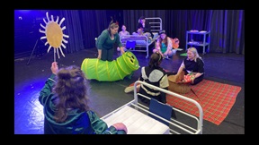 Drama students on stage in hospital set in various poses, one moving a large model Hungry Caterpillar
