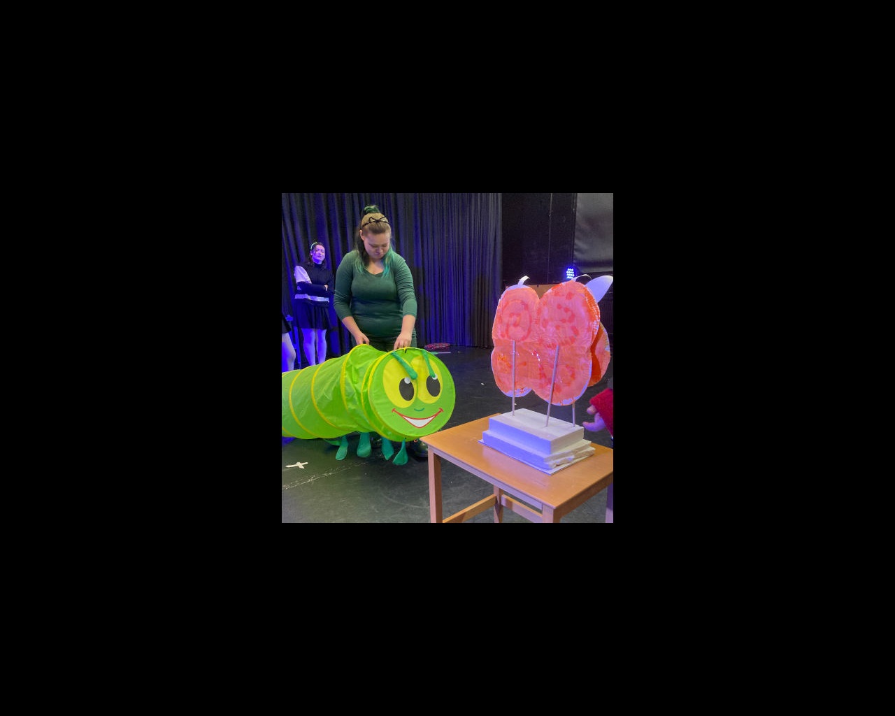 Female student holding large model Hungry Caterpillar up to table with butterfly-like model on it.