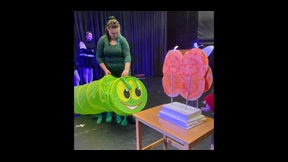 Female student holding large model Hungry Caterpillar up to table with butterfly-like model on it.
