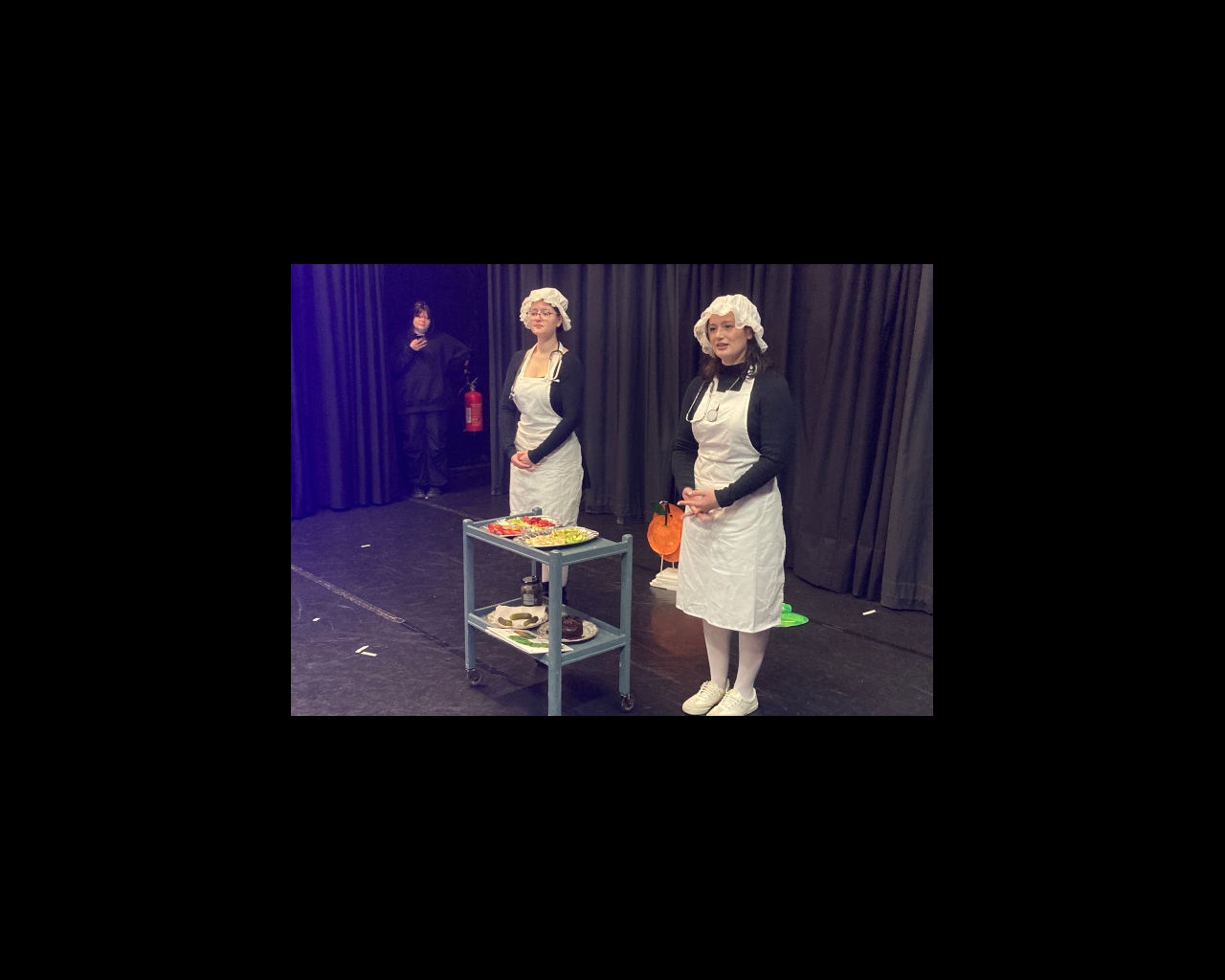 Two female drama students dressed in old-fashioned nurse uniforms with trolley of food on stage